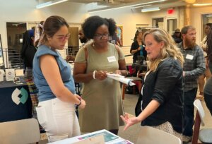 A woman pointing to a flyer on a table. Two other women are standing next to her, looking at the flyer.